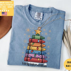 以下が含まれることがあります： 青いジーンズカラーのTシャツ。クレヨンで作られたクリスマスツリーのデザイン。ツリーは様々な色のクレヨンを積み重ねて作られており、上には星、下には「Early Intervention」の文字。金のネックレスがシャツの上にかけられています。
