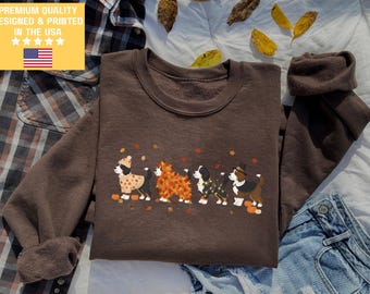 Puede incluir: Una sudadera marrón con un diseño de perros con disfraces otoñales. Los perros están decorados con hojas y calabazas. El texto "PREMIUM QUALITY DESIGNED & PRINTED IN THE USA" está en un cuadro amarillo con la bandera estadounidense.