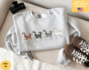 Op de afbeelding: Lichtgrijs sweatshirt met een rij van vijf cartoon husky's in verschillende kleuren. De sweatshirt heeft een ronde hals en lange mouwen. De tekst "PREMIUM QUALITY DESIGNED & PRINTED IN THE USA - SHIPS FAST!" is gedrukt op een gele achtergrond.