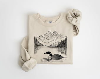 May include: Beige sweatshirt featuring a black and white illustration of a mountain range, lake, and a loon. The loon is swimming in the water. The sweatshirt has a crew neck and long sleeves. The sleeves are rolled up.