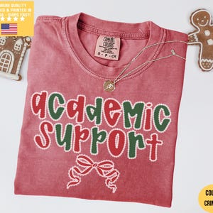 Può includere: T-shirt color cremisi con le parole "academic support" in rosso e verde, con una grafica a fiocco. La maglietta ha uno scollo tondo e un'etichetta Comfort Colors. Una collana d'argento con un ciondolo è sulla maglietta. Un omino di pan di zenzero e una casa sono sullo sfondo.