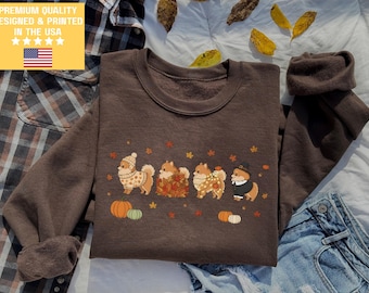 May include: A brown sweatshirt featuring a fall-themed design with four Pomeranian dogs dressed in autumn attire. The dogs are surrounded by pumpkins and fall leaves. The text "PREMIUM QUALITY DESIGNED & PRINTED IN THE USA" is in the upper left corner.