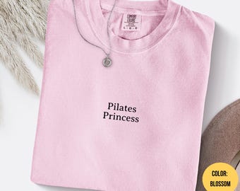 Puede incluir: Una camiseta rosa claro con la frase "Pilates Princess" en negro. Un collar plateado con un colgante circular descansa sobre la camiseta. La camiseta está doblada y tiene una etiqueta "Comfort Colors".