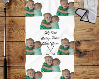 May include: White golf towel featuring multiple images of two young boys hugging. The towel has the text "My Dad Swings Better Than Yours" with crossed golf clubs. A golf club, glove, and ball are also visible.