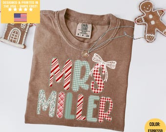 May include: Espresso-colored t-shirt with the text "MRS MILLER" in a festive design. The letters feature candy cane stripes, gingham, and a bow. A silver necklace with a pendant is visible. A gingerbread man and house are in the background.
