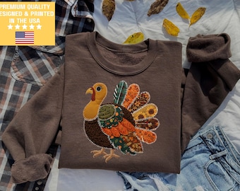 Op de afbeelding: Een bruine sweatshirt met een kleurrijk kalkoenontwerp. De kalkoen heeft een gele kop, een rode lellen en een patchwork van oranje, groene en bruine veren. De tekst "PREMIUM QUALITY DESIGNED & PRINTED IN THE USA" staat linksboven.