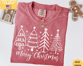 May include: Crimson-colored Christmas t-shirt with a white graphic of stylized Christmas trees and the words "Merry Christmas." The shirt has a crew neck and short sleeves. A silver necklace with a pendant is on the shirt.