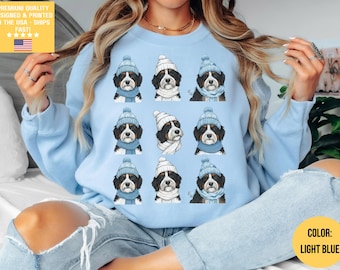 Puede incluir: Sudadera azul claro con un estampado repetido de perros de montaña berneses con gorros y bufandas de invierno. Los perros están representados en un diseño repetitivo. El texto "PREMIUM QUALITY DESIGNED & PRINTED IN THE USA - SHIPS FAST!" está en la esquina superior izquierda.