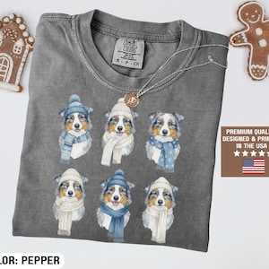 May include: A pepper-coloured t-shirt with a crew neck, displaying a watercolour illustration of six Australian Shepherds in winter attire. The shirt has a tag that reads "COMFORT COLORS". A gold necklace and gingerbread cookies are also visible.