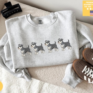 Può includere: Felpa color cenere con una fila di illustrazioni di cani Schnauzer. La felpa ha un girocollo e maniche lunghe. L'immagine mostra anche scarpe da ginnastica marroni con strisce bianche e il testo "PREMIUM QUALITY DESIGNED & PRINTED IN THE USA - SHIPS FAST!"