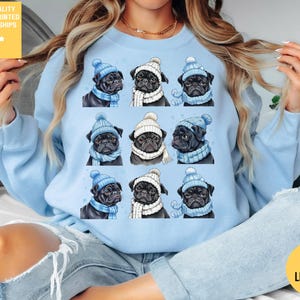 Könnte beinhalten: Hellblaues Sweatshirt mit einem Muster aus neun Möpsen, die Wintermützen und Schals tragen. Einige Möpse haben blaue Mützen und Schals, andere weiße. Der Text "PREMIUM QUALITY DESIGNED & PRINTED IN THE USA - SHIPS FAST!" befindet sich oben links.