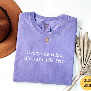 Könnte beinhalten: Violettes Comfort Colors T-Shirt mit dem Text "Everyone relax, It's our Girls Trip." Eine goldene Halskette mit Anhänger liegt auf dem Shirt. Ein brauner Hut und getrocknete Palmblätter sind ebenfalls im Bild.