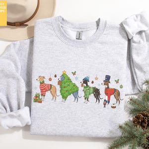 Könnte beinhalten: Hellgraues Sweatshirt mit einem weihnachtlichen Design. Die Grafik zeigt vier Hunde in festlichen Outfits, darunter einen rot-weiß gestreiften Pullover und ein Weihnachtsbaumkostüm. Das Sweatshirt hat einen Rundhalsausschnitt und lange Ärmel.