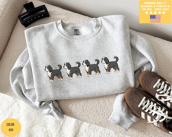Puede incluir: Sudadera gris ceniza con un diseño bordado de cinco perros de montaña berneses. Los perros están representados en fila, con marcas negras, blancas y marrones. La sudadera se combina con zapatillas marrones. El texto "PREMIUM QUALITY DESIGNED & PRINTED IN THE USA - SHIPS FAST!" es visible.