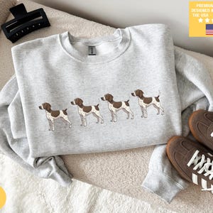Op de afbeelding: Asgrijze sweatshirt met een geborduurd ontwerp van vier bruine en witte honden. De sweatshirt heeft een ronde hals en lange mouwen. De afbeelding toont ook bruine sneakers, een zwarte haarclip en een gele sticker met tekst.