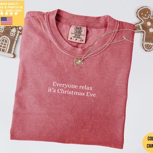May include: Crimson t-shirt with the text "Everyone relax it's Christmas Eve." A silver necklace with a pendant is on the shirt. A gingerbread man and house are in the background.