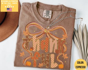 May include: Espresso-colored t-shirt with the word "thankful" in a fall-themed design. The design includes a large bow, pumpkins, and leaves. A silver necklace with a small pendant is on the shirt.