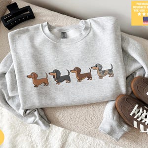 Könnte beinhalten: Hellgraues Sweatshirt mit einer Reihe von vier Dackel-Illustrationen in verschiedenen Farben. Das Sweatshirt hat einen Rundhalsausschnitt und lange Ärmel. Der Text "PREMIUM QUALITY DESIGNED & PRINTED IN THE USA - SHIPS FAST!" ist auf gelbem Hintergrund gedruckt.