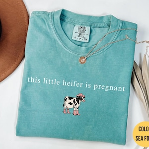 Könnte beinhalten: Ein kurzärmeliges T-Shirt in Meerschaumgrün mit Rundhalsausschnitt. Das Shirt zeigt den weißen Schriftzug "this little heifer is pregnant" über einer Grafik einer schwarz-weißen Kuh mit einem rosa Cowboyhut und rosa Cowboystiefeln.