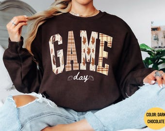 May include: A dark chocolate brown crewneck sweatshirt featuring the word GAME in large block letters with a tan plaid and autumn leaf pattern. Below the word GAME, the word day is printed in white script, flanked by two small white football icons.
