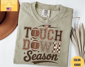 May include: Khaki-colored t-shirt with the words "TOUCH DOWN SEASON" in brown lettering. The design includes footballs and a mix of patterns. The shirt has a Comfort Colors tag and a small necklace. The color is khaki.