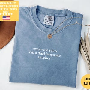May include: Blue jean colored t-shirt with the text "everyone relax I'm a dual language teacher" in white. A gold necklace with a pendant rests on the shirt. The shirt has a Comfort Colors tag.