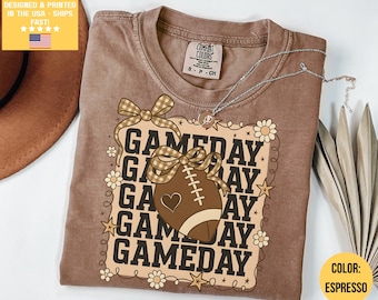 May include: Espresso-colored t-shirt with a 'GAMEDAY' graphic featuring a football, bows, and floral accents. The shirt has a crew neck and short sleeves. A silver necklace with a pendant is draped on the shirt.
