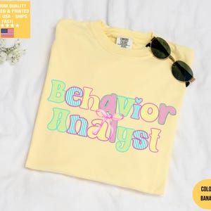 Könnte beinhalten: Hellgelbes T-Shirt mit dem Schriftzug "Behavior Analyst" in bunten, gestreiften Buchstaben. Das T-Shirt hat einen Rundhalsausschnitt und eine lockere Passform. Eine Sonnenbrille liegt auf dem T-Shirt. Das Shirt ist aus weichem Material.