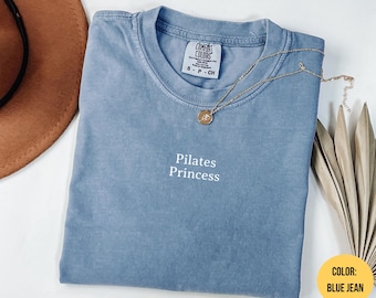Puede incluir: Una camiseta azul vaquero con las palabras "Pilates Princess" en blanco. La camiseta tiene cuello redondo y una pequeña etiqueta en el cuello. Un collar dorado con un colgante está drapeado sobre la camiseta.