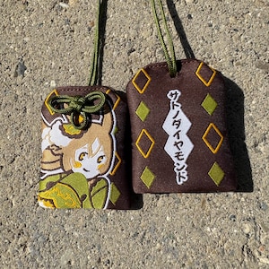 May include: Two brown fabric omamori charms with green cords. One features an embroidered anime-style character in green and gold clothing. The other displays yellow diamond shapes and white Japanese text reading Satono Diamond.