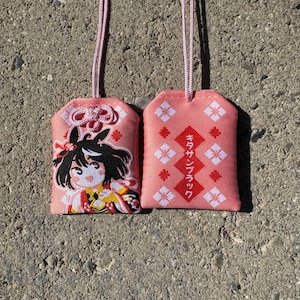 May include: Two rectangular pink fabric omamori charms with light pink hanging cords. One charm features an embroidered anime-style character with black hair, while the other displays red diamond shapes and Japanese text reading キタサンブラック.