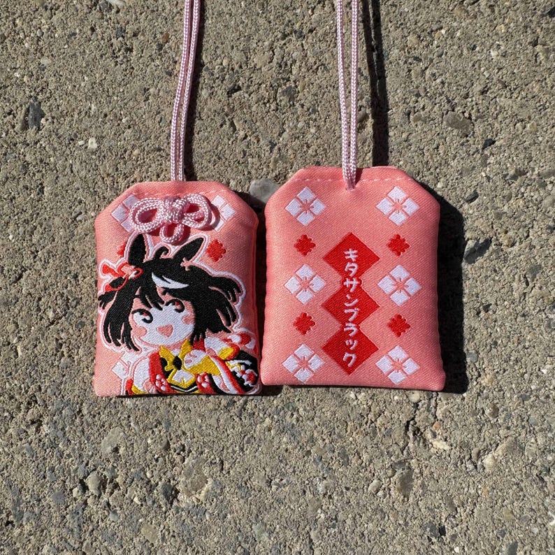 May include: Two rectangular pink fabric omamori charms with light pink hanging cords. One charm features an embroidered anime-style character with black hair, while the other displays red diamond shapes and Japanese text reading キタサンブラック.