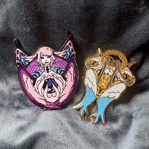 May include: Two enamel pins featuring stylized figures. One pin has a pink and purple design with a central figure and outstretched wings. The other pin has a gold and white design with blue accents, also depicting a figure with wings.