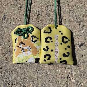 May include: Two yellow fabric omamori charms with a black leopard print pattern. One charm features an embroidered anime-style character with brown hair, while the other displays vertical Japanese text reading Jungle Pocket.