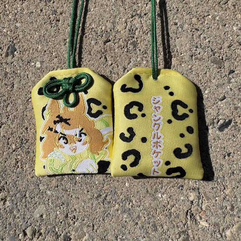 May include: Two yellow fabric omamori charms with a black leopard print pattern. One charm features an embroidered anime-style character with brown hair, while the other displays vertical Japanese text reading Jungle Pocket.