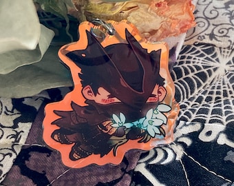 May include: A black and orange anime-style character with a mask and flowers. The character is holding a bouquet of white flowers. The character is on a clear acrylic keychain.