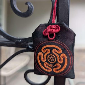 Puede incluir: Un amuleto de tela negra con un diseño circular naranja y marrón. El amuleto está suspendido por un cordón y un nudo rojos, colgando de una estructura metálica negra. El diseño presenta un patrón estilizado.