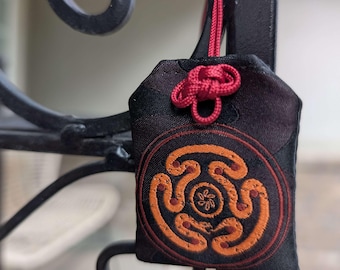 May include: A black fabric charm with an orange and brown circular design. The charm is suspended by a red cord and knot, hanging from a black metal structure. The design features a stylized pattern.