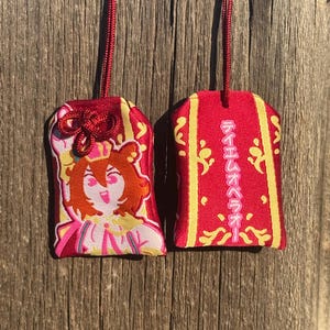 May include: Two rectangular red fabric omamori charms with gold trim and red hanging cords. One charm features an embroidered anime-style character with brown hair and a crown. The other charm displays vertical Japanese text in pink, reading Teiemu Opera O, surrounded by gold decorative patterns.