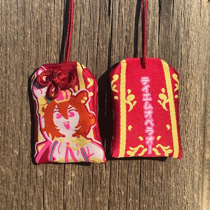 May include: Two rectangular red fabric omamori charms with gold trim and red hanging cords. One charm features an embroidered anime-style character with brown hair and a crown. The other charm displays vertical Japanese text in pink, reading Teiemu Opera O, surrounded by gold decorative patterns.