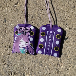 May include: Two purple fabric omamori charms with white and black polka dots and lavender hanging cords. One charm features an embroidered anime-style character with purple hair, while the other displays the Japanese text メジロマックイーン.