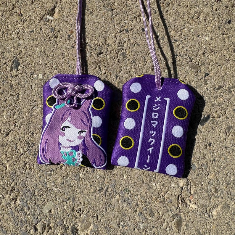 May include: Two purple fabric omamori charms with white and black polka dots and lavender hanging cords. One charm features an embroidered anime-style character with purple hair, while the other displays the Japanese text メジロマックイーン.