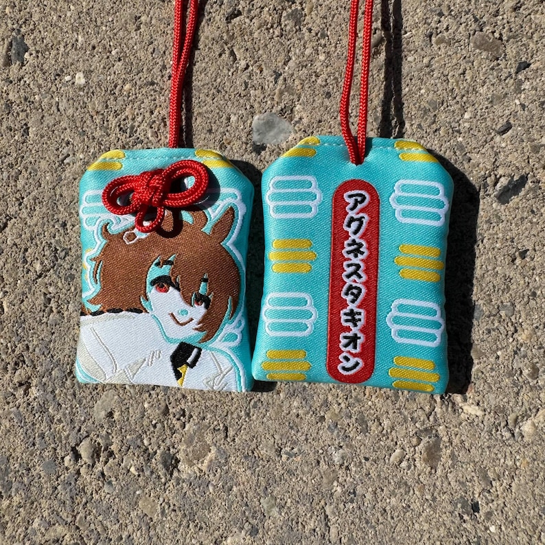 May include: Two rectangular omamori charms in light blue fabric with red hanging cords. One charm features an embroidered anime-style character with brown hair, while the other displays a vertical red label with Japanese text and yellow accents.