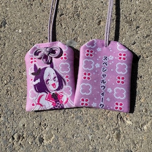 May include: Two rectangular lavender fabric omamori charms with matching cords. One charm features an embroidered anime-style character with dark hair, while the other displays white clover patterns and Japanese text reading スペシャルウィーク.