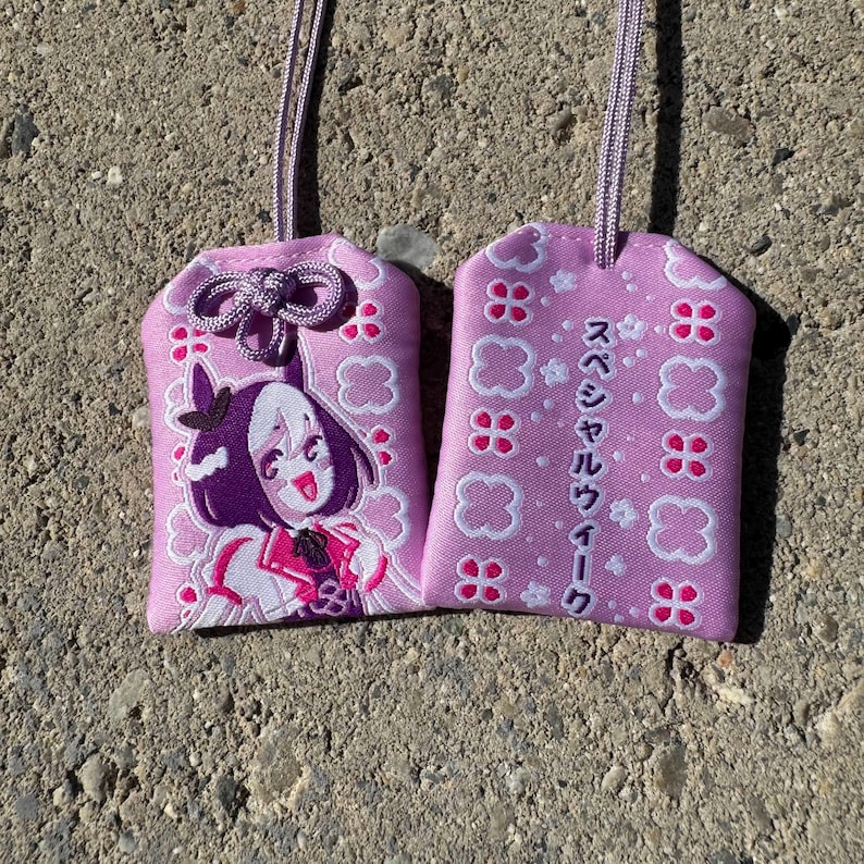May include: Two rectangular lavender fabric omamori charms with matching cords. One charm features an embroidered anime-style character with dark hair, while the other displays white clover patterns and Japanese text reading スペシャルウィーク.