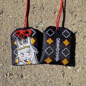 May include: Two black fabric omamori charms with red hanging cords. One charm features an embroidered white anime-style character with a yellow headband. The other charm displays yellow and white diamond patterns and Japanese text in white embroidery.