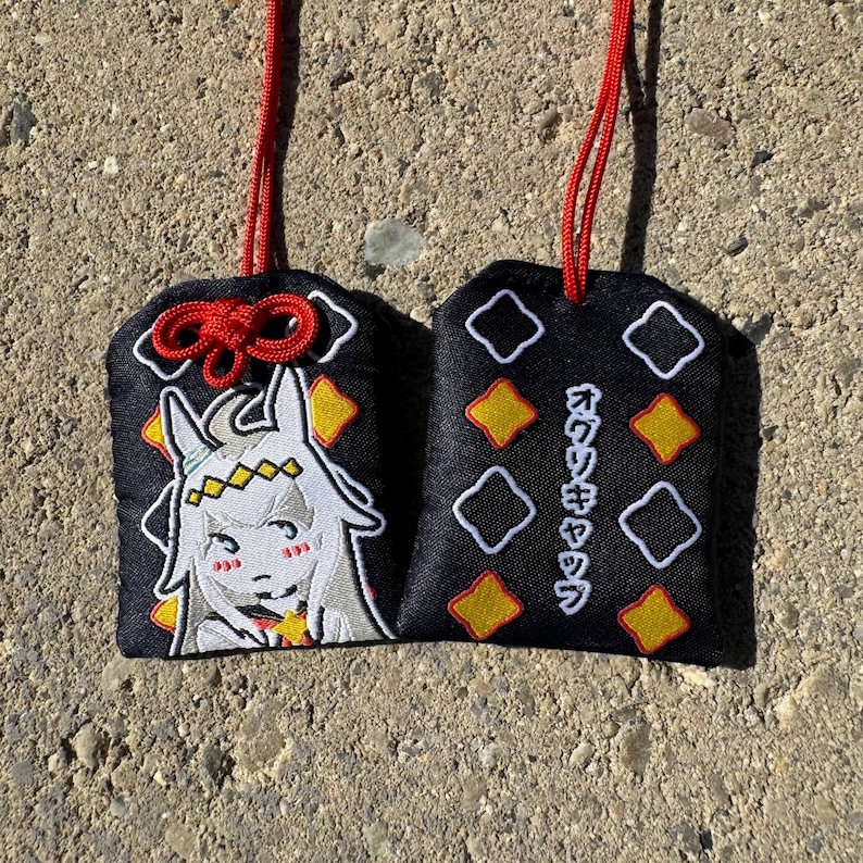 May include: Two black fabric omamori charms with red hanging cords. One charm features an embroidered white anime-style character with a yellow headband. The other charm displays yellow and white diamond patterns and Japanese text in white embroidery.