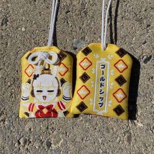 May include: Two yellow fabric omamori charms with white hanging cords. One charm features an embroidered white rabbit character with red accents. The other charm displays a vertical white rectangular border containing the Japanese text Gold Ship.