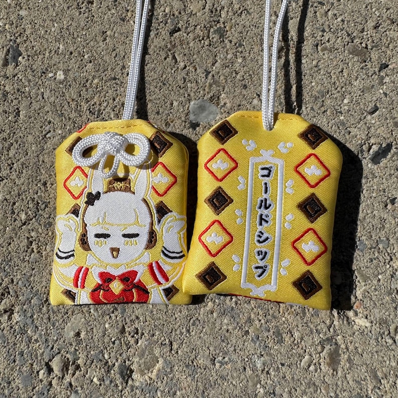 May include: Two yellow fabric omamori charms with white hanging cords. One charm features an embroidered white rabbit character with red accents. The other charm displays a vertical white rectangular border containing the Japanese text Gold Ship.