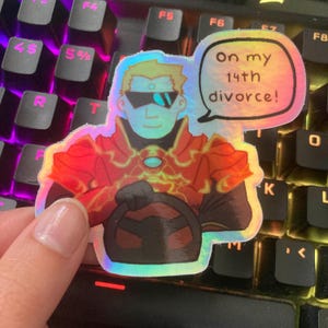 Op de afbeelding: Een holografische sticker met een cartoonfiguur dat een zonnebril en een rood-zwarte outfit draagt. Het figuur houdt een stuurwiel vast. De sticker heeft een spraakballon met de tekst "On my 14th divorce!"